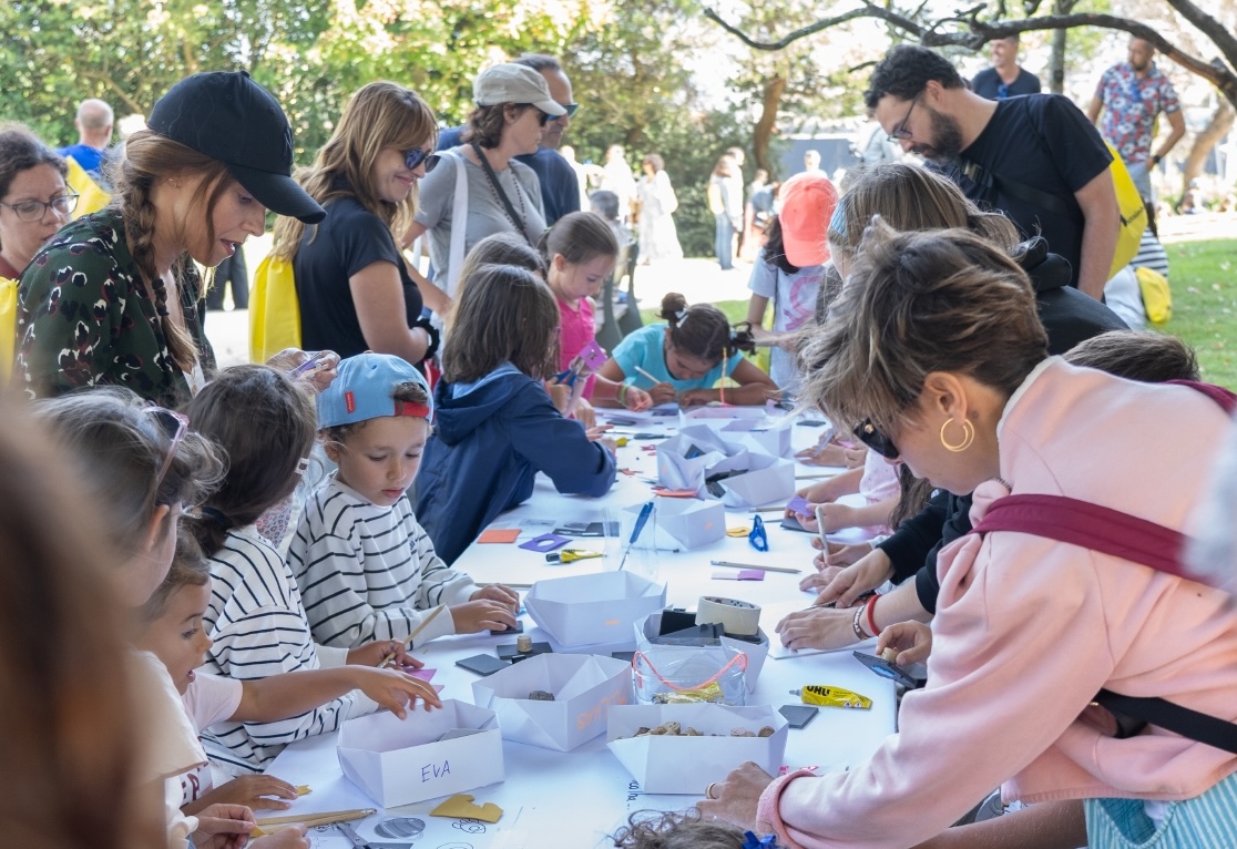 Grupo de crianças e adultos num jardim, à volta de uma mesa, a participar numa atividade criativa durante um evento, simbolizando a cobertura integral de eventos da 4 Ventos que capta momentos autênticos, interação e envolvimento do público.